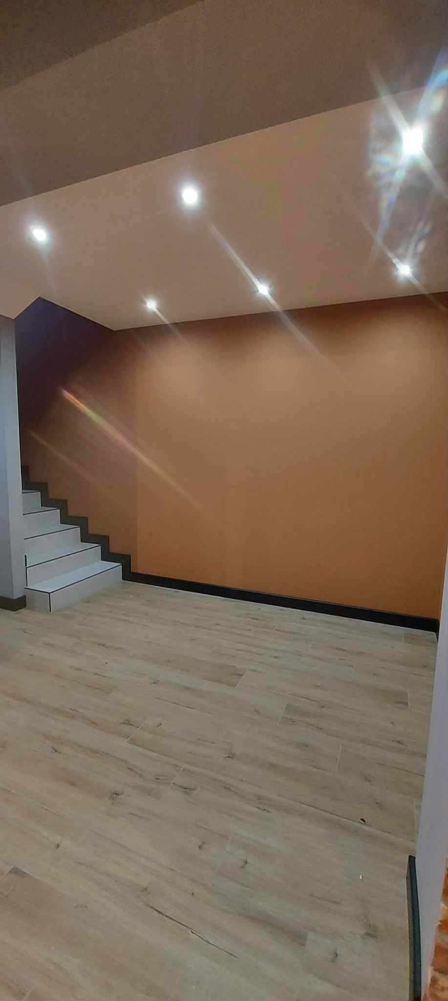 Beżowa ściana z oświetleniem punktowym i drewnopodobna podłoga. Widoczne schody z szarymi stopniami i czarną listwą przypodłogową. Ukończone wnętrze po remoncie.