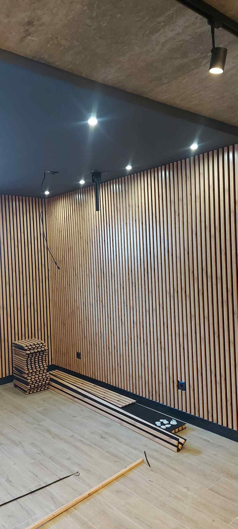 Montaż drewnianych lameli na ścianie z oświetleniem punktowym. Część lameli ułożona na podłodze. Widoczne gniazdka elektryczne. Surowy beton na suficie.