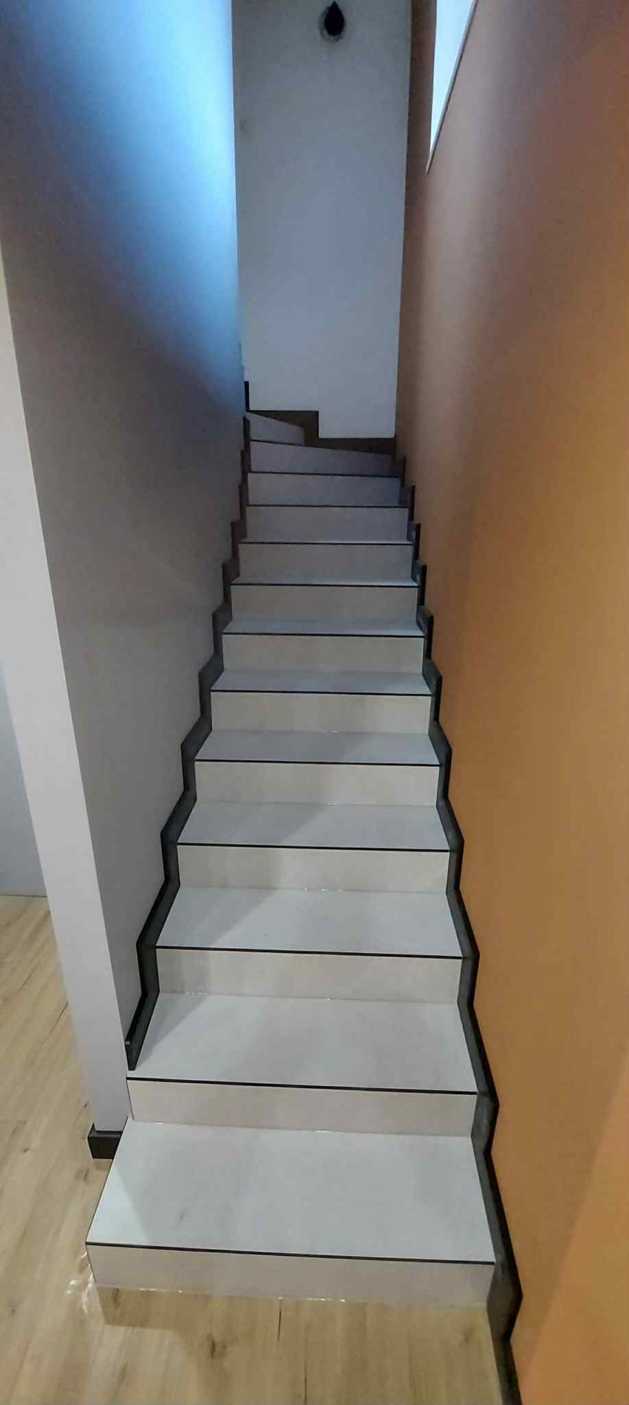 Białe schody z czarnymi krawędziami w korytarzu z szarą i pomarańczową ścianą. Widok z dołu, oświetlenie górne.