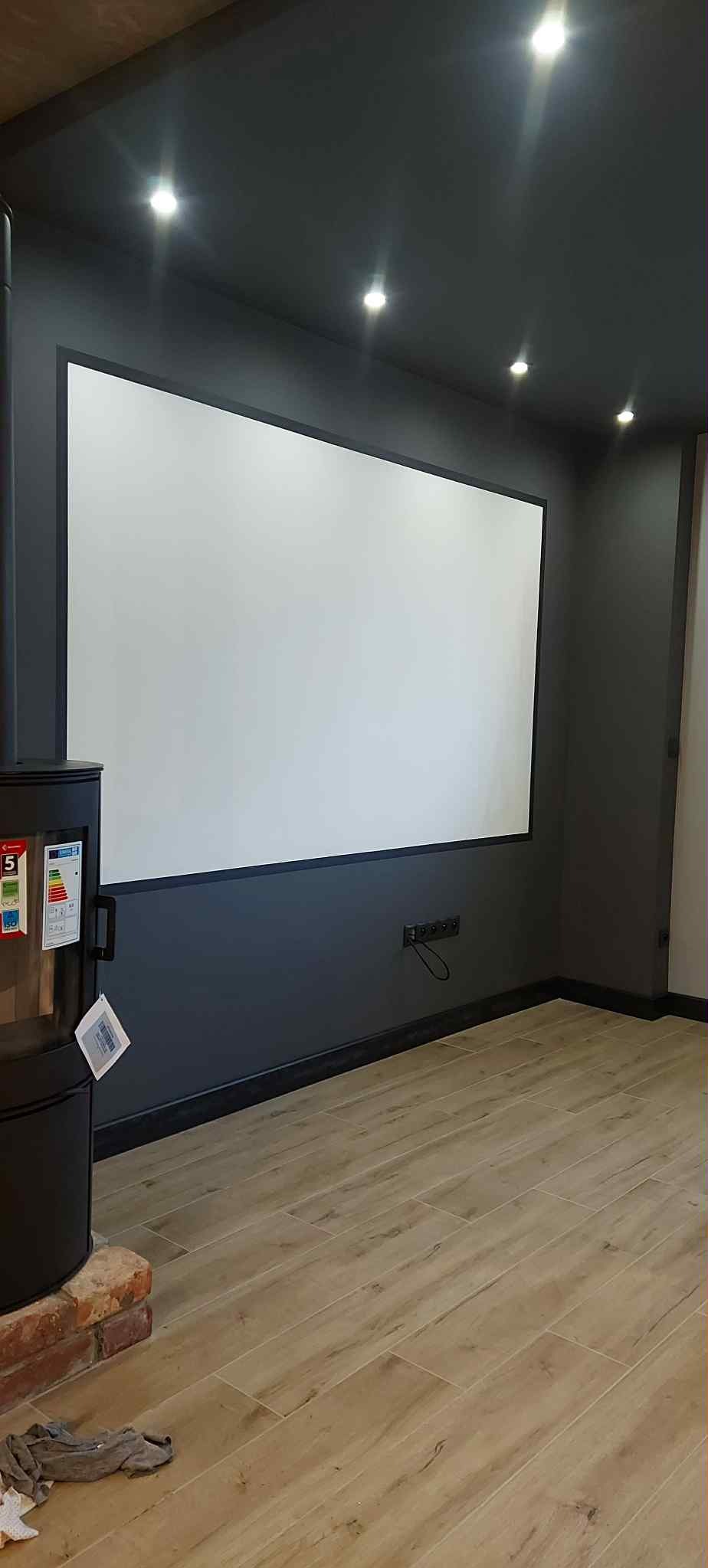 Nowoczesne wnętrze z ekranem projekcyjnym na ścianie, drewnopodobna podłoga, ciemnoszara ściana z oświetleniem punktowym i piec wolnostojący z boku.