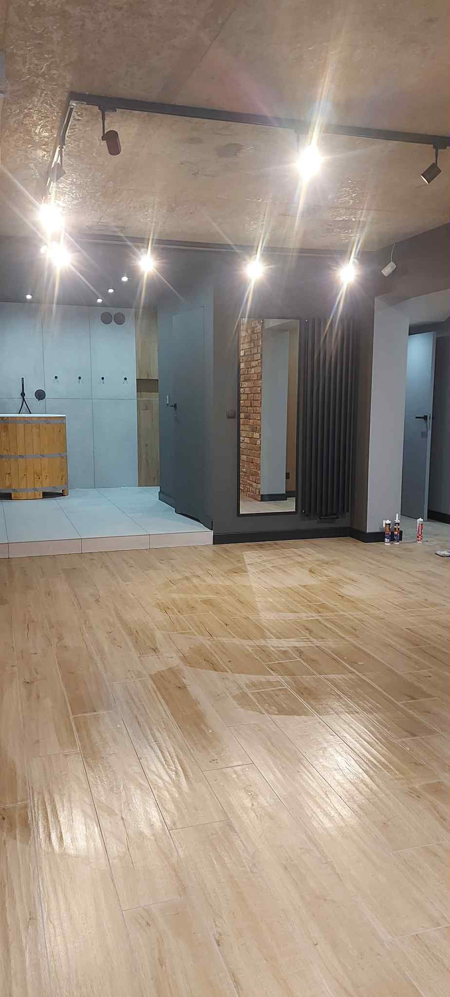 Jasne panele podłogowe w wykończonym wnętrzu z szarą zabudową i oświetleniem punktowym. Widoczne akcesoria montażowe w tle.