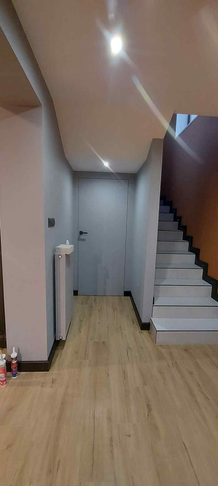 Jasne panele podłogowe w korytarzu z szarą ścianą, grzejnikiem i drzwiami. Schody z białymi stopniami i pomarańczową ścianą w tle. Nowoczesne wykończenie.