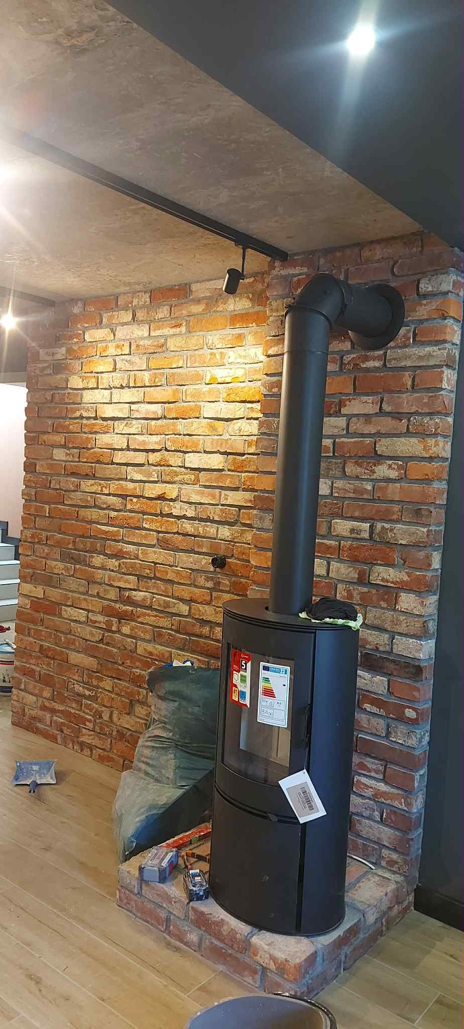 Ceglana ściana z kominem w salonie, oświetlenie szynowe na suficie. Widoczne narzędzia i worki, co sugeruje etap wykończeniowy. Parkiet na podłodze.