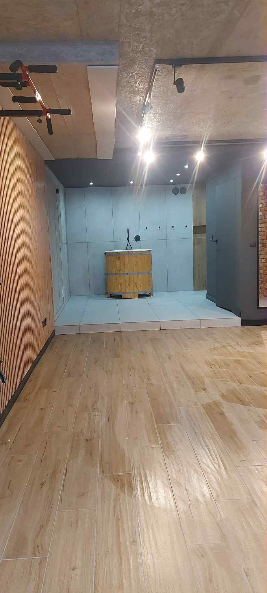 Nowoczesne wnętrze z drewnianą podłogą, panelami ściennymi i podwyższoną platformą z drewnianą beczką. Oświetlenie punktowe i surowy betonowy sufit.