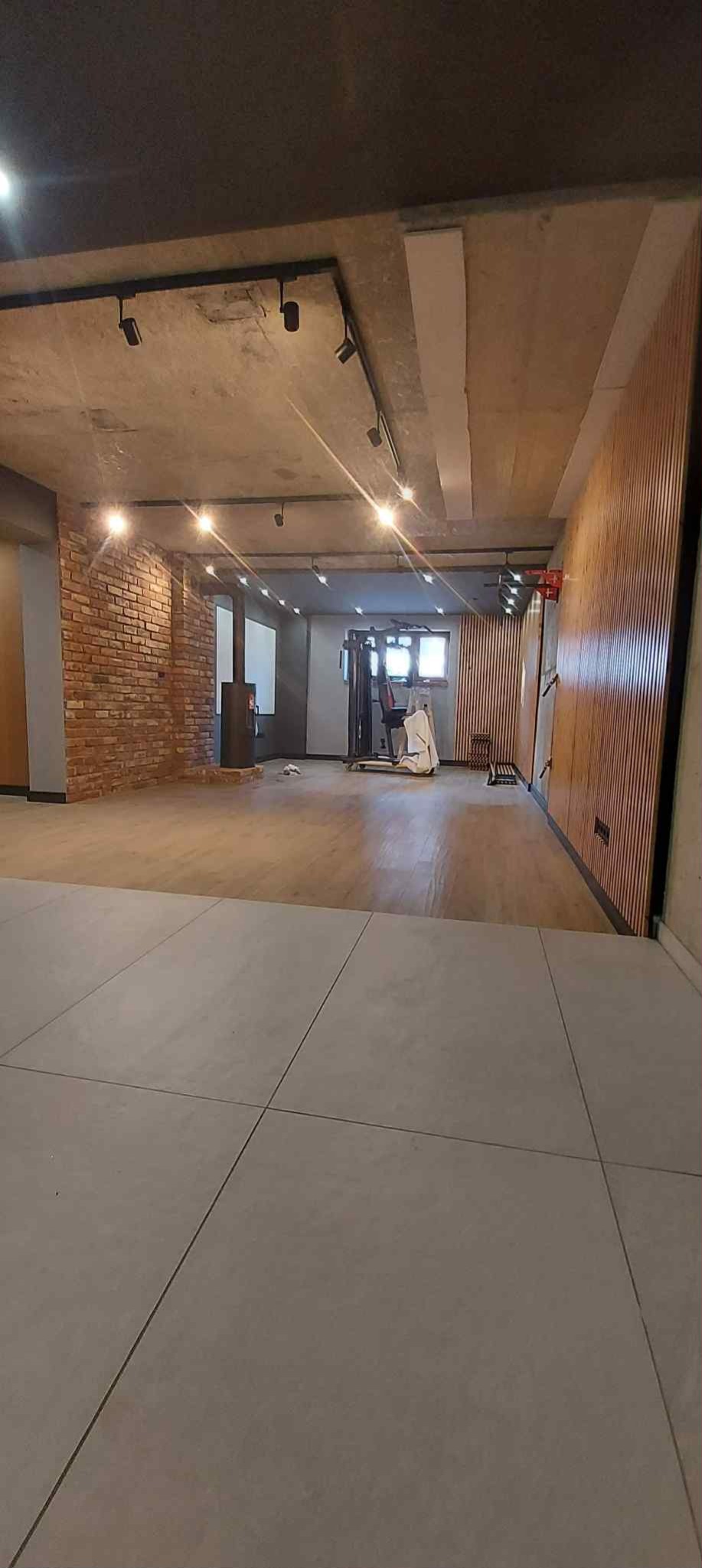 Przestronne wnętrze po remoncie: drewniana podłoga, ceglana ściana, panele ścienne, nowoczesne oświetlenie punktowe i szare płytki podłogowe. Widok na siłownię.