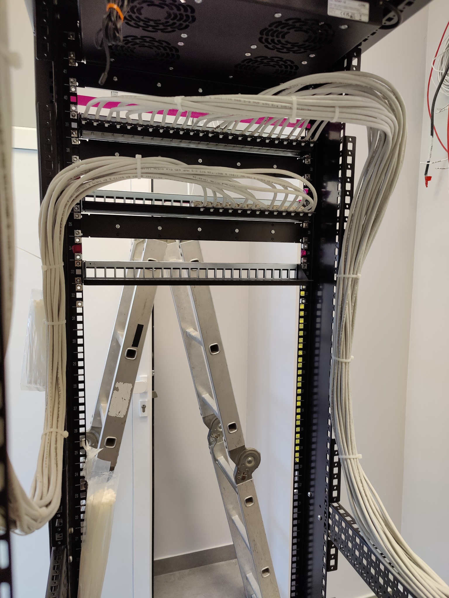 Serwerownia w trakcie instalacji okablowania sieciowego, widoczna drabina oparta o szafę rack, przewody uporządkowane w wiązki, panele krosownicze częściowo podłączone.
