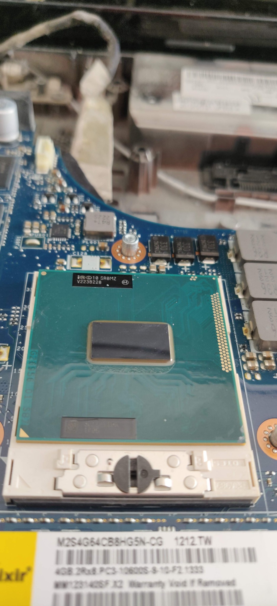 Otwarta obudowa laptopa z widocznym procesorem z nałożoną pastą termoprzewodzącą, niebieska płyta główna z elementami elektronicznymi, widoczne złącze RAM i częściowo etykieta z kodem kreskowym.