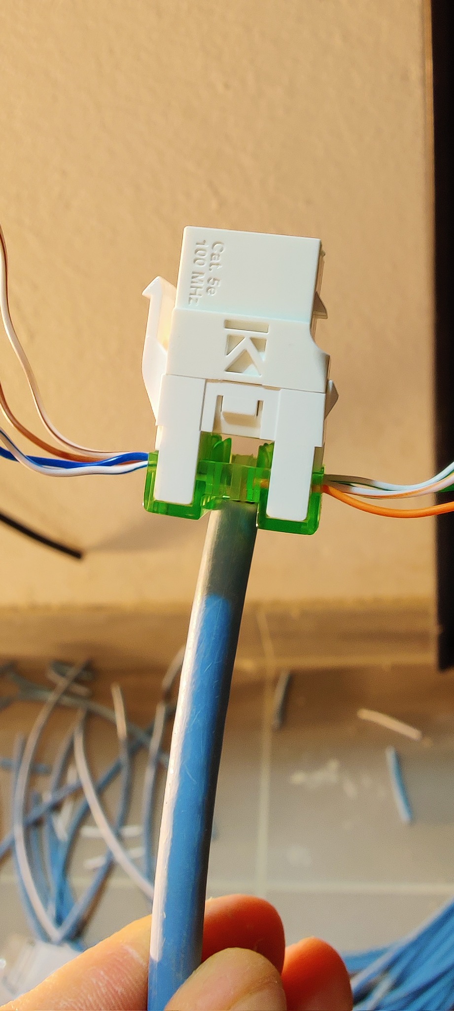 Złącze Cat5e 100 MHz z zielonymi elementami mocującymi, podłączone do niebieskiego kabla sieciowego, w tle fragmenty kabli.