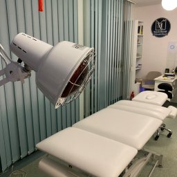 Małgorzata Basińska Gabinet Masażu MusculeMassage - Biały stół do zabiegów z lampą grzewczą w gabinecie, zasłony pionowe w tle. Widoczne wyposażenie i logo firmy na ścianie. Wnętrze gabinetu.