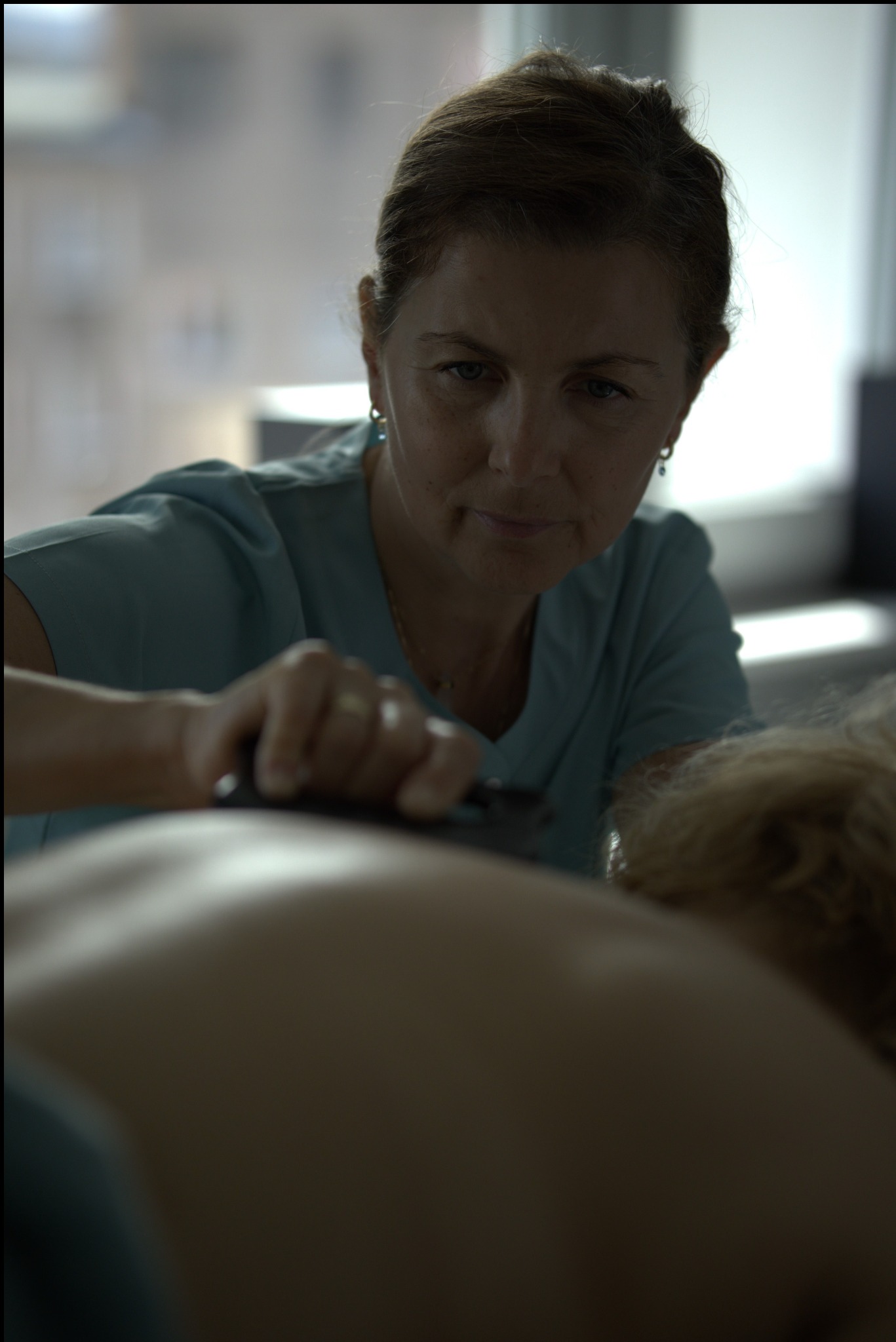 Kobieta w turkusowej koszulce wykonuje zabieg na plecach klienta przy użyciu czarnego narzędzia. Delikatne oświetlenie, skupienie na dłoniach i plecach.