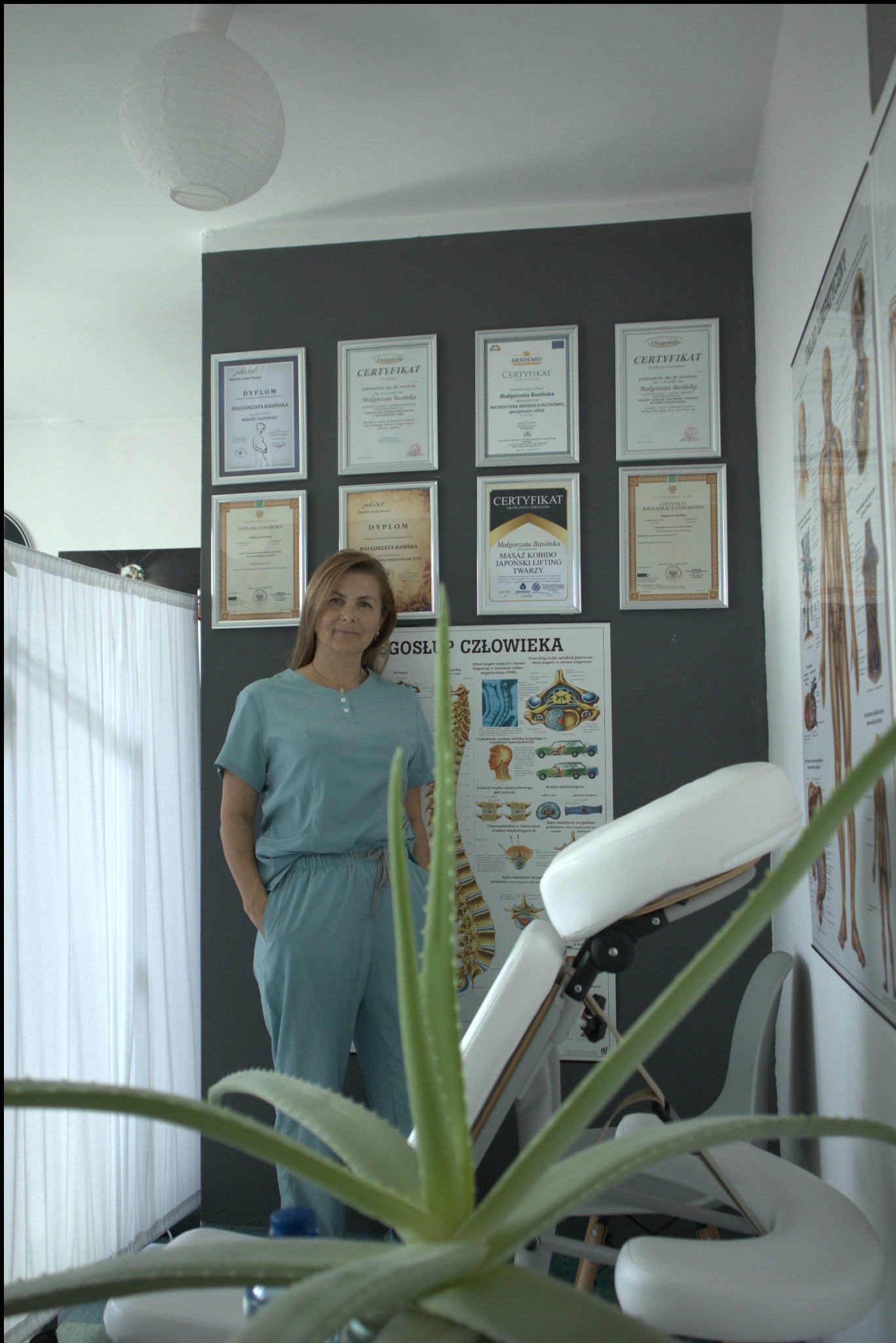 Gabinet z certyfikatami masażu, tablicą anatomiczną i fotelem. Uśmiechnięta terapeutka w zielonym uniformie pozuje, z przodu roślina doniczkowa.