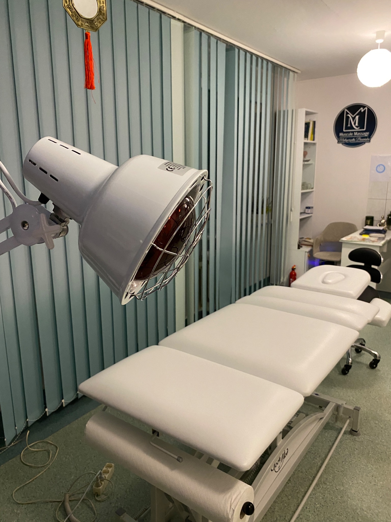 Biały stół do zabiegów z lampą grzewczą w gabinecie, zasłony pionowe w tle. Widoczne wyposażenie i logo firmy na ścianie. Wnętrze gabinetu.