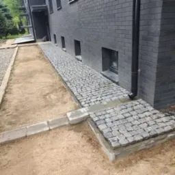 Świeżo ułożona ścieżka z kostki granitowej prowadząca wzdłuż ciemnej, ceglanej ściany budynku, widoczne niedokończone obrzeża i przygotowane podłoże.