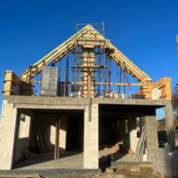 Budowa domu w stanie surowym otwartym: ściany z bloczków, drewniane szalunki dachu, podpory montażowe i rusztowanie na tle błękitnego nieba.