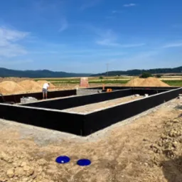 Budowa fundamentów z bloczków betonowych pomalowanych na czarno, dwóch pracowników w trakcie prac ziemnych, widok na pagórkowaty krajobraz w tle.