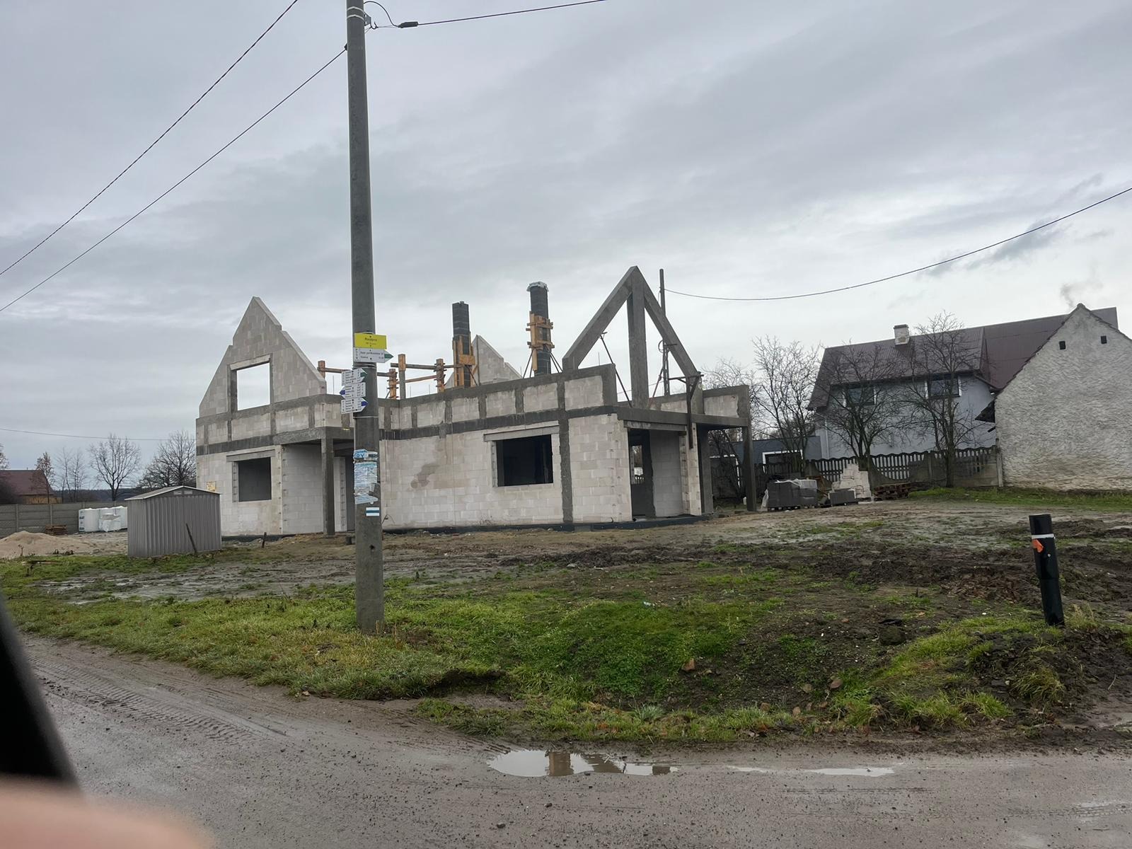 Surowy stan domu murowanego w trakcie budowy, widoczne ściany z bloczków betonowych, drewniane szalunki kominowe, słup elektryczny z ogłoszeniami na pierwszym planie, błotniste podłoże.