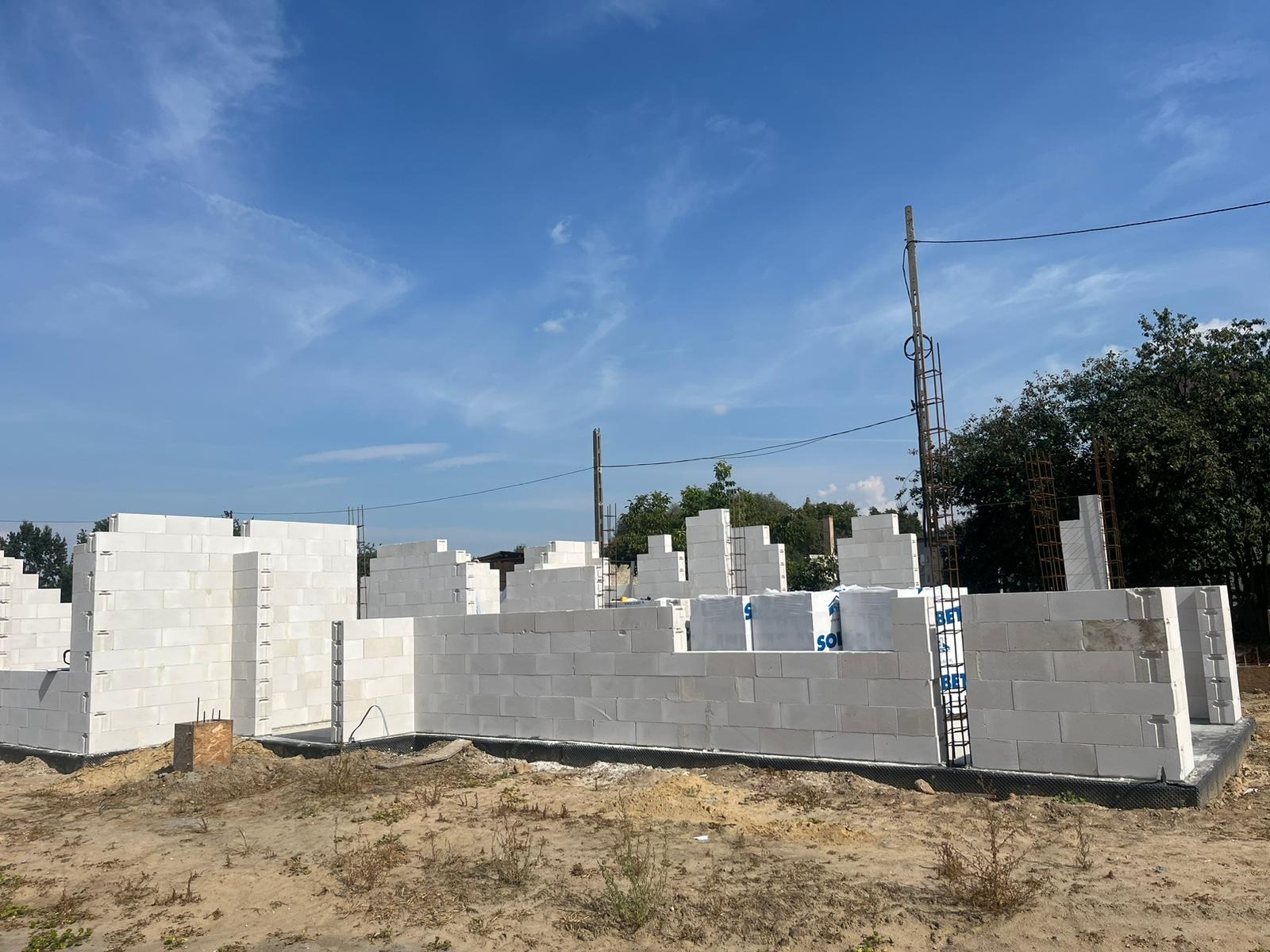 Wznoszenie ścian parteru z bloczków betonu komórkowego na fundamencie, widoczne zbrojenia pionowe i poziome, w tle niebo i drzewa.