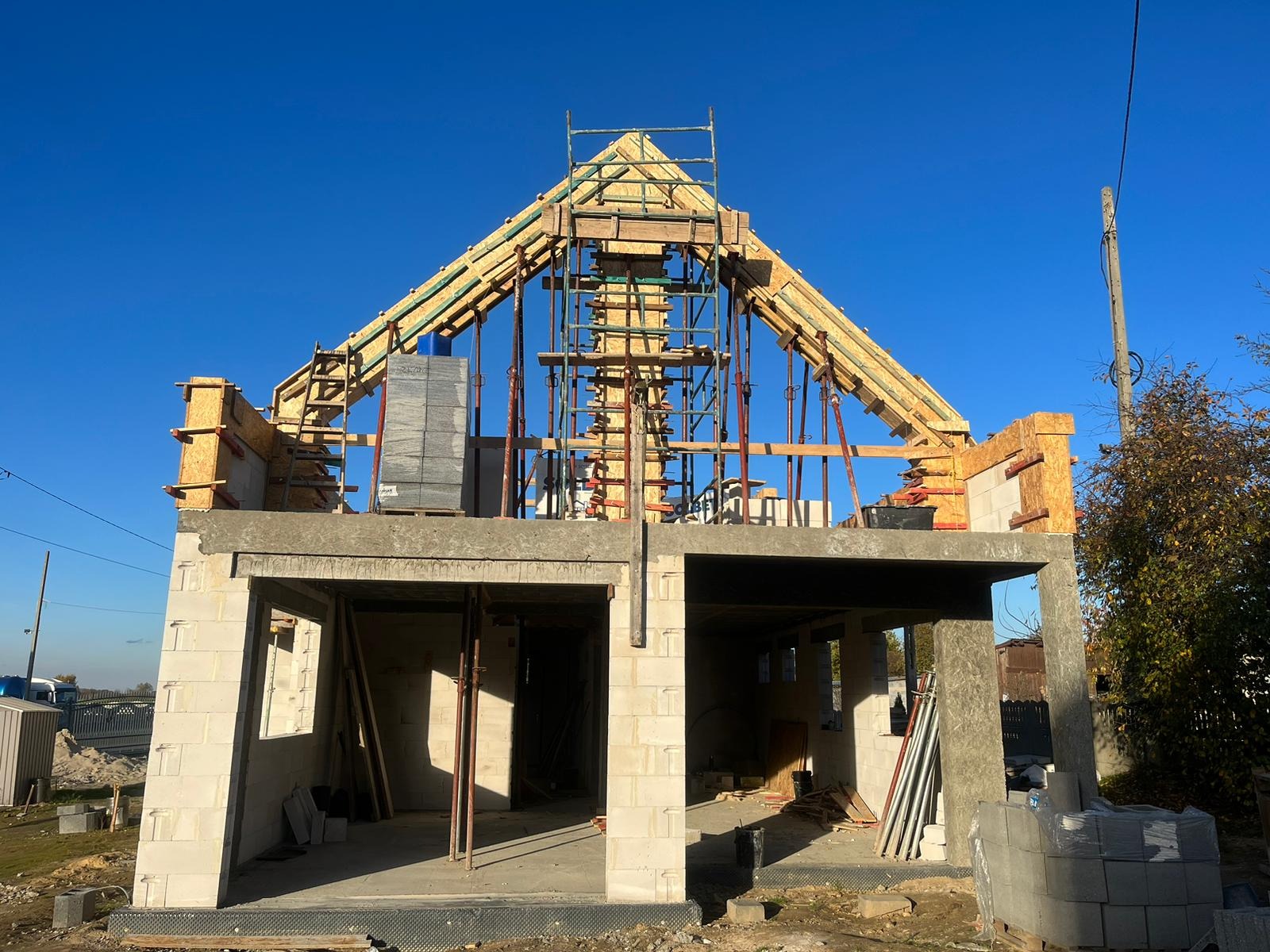 Budowa domu w stanie surowym otwartym: ściany z bloczków, drewniane szalunki dachu, podpory montażowe i rusztowanie na tle błękitnego nieba.