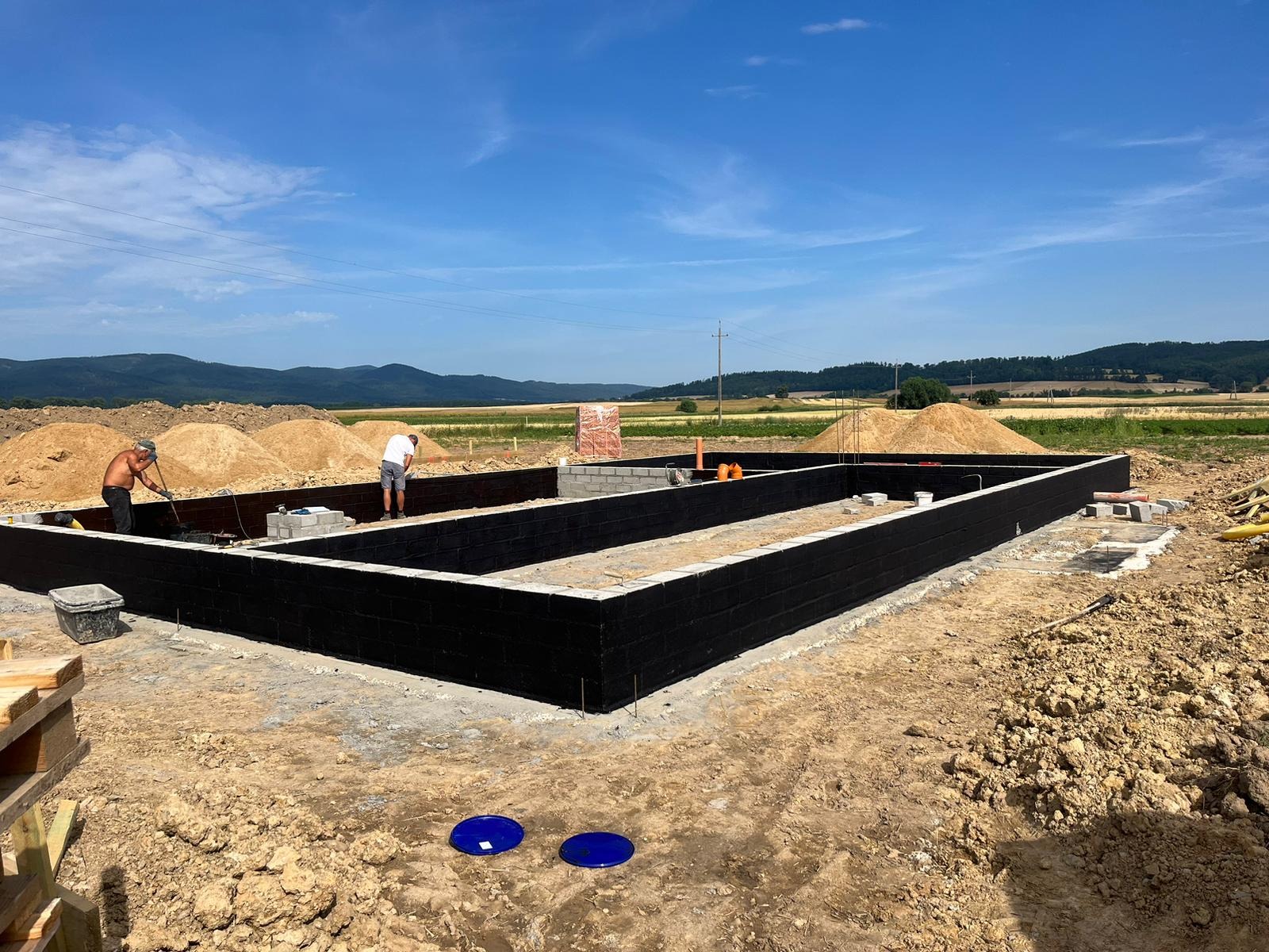 Budowa fundamentów z bloczków betonowych pomalowanych na czarno, dwóch pracowników w trakcie prac ziemnych, widok na pagórkowaty krajobraz w tle.