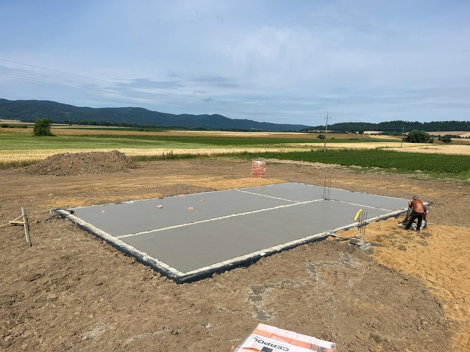 Wylany betonowy fundament na działce z widokiem na pola i wzgórza, widoczny pracownik przy pracy, wystające pręty zbrojeniowe i cegły w tle.