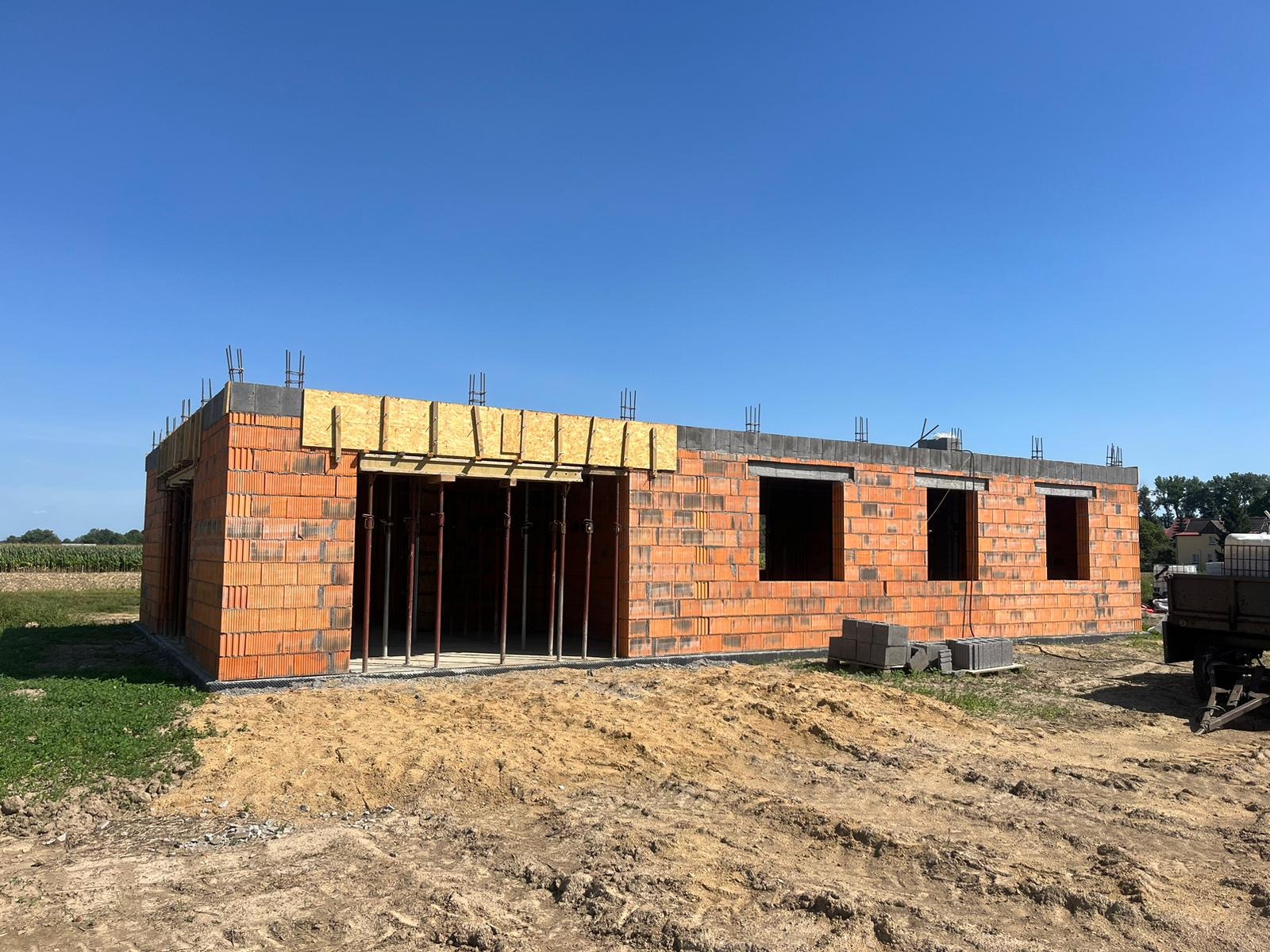 Wznoszenie ścian parterowego budynku z czerwonej cegły, widoczne zbrojenie i drewniane szalunki, teren budowy z piaskiem i częściowo zarośnięty trawą, błękitne niebo w tle.