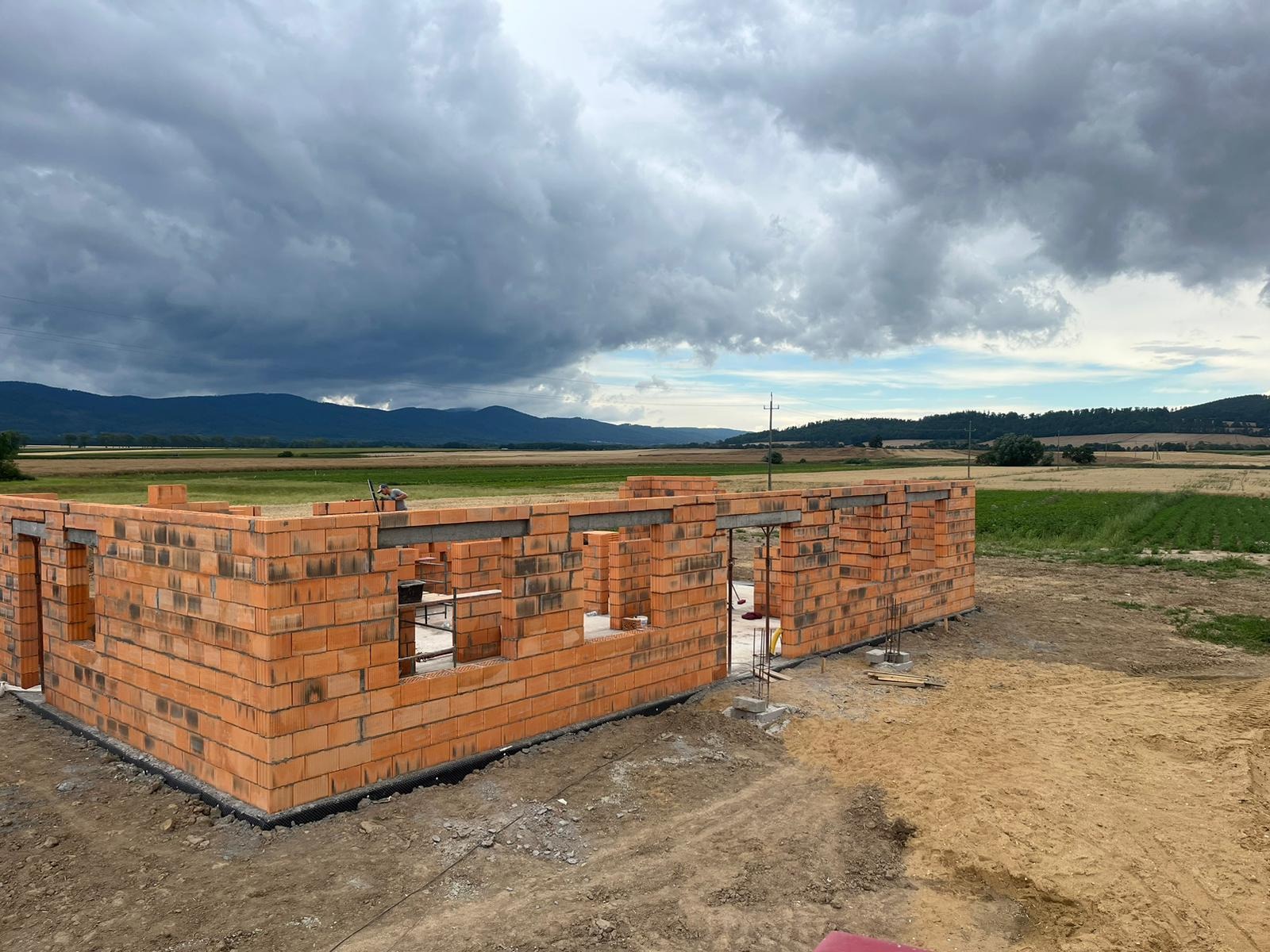 Wznoszone ściany parteru domu z czerwonej cegły, widoczne stalowe nadproża okienne i drzwiowe, w tle krajobraz pól i zalesionych wzgórz pod burzowym niebem.