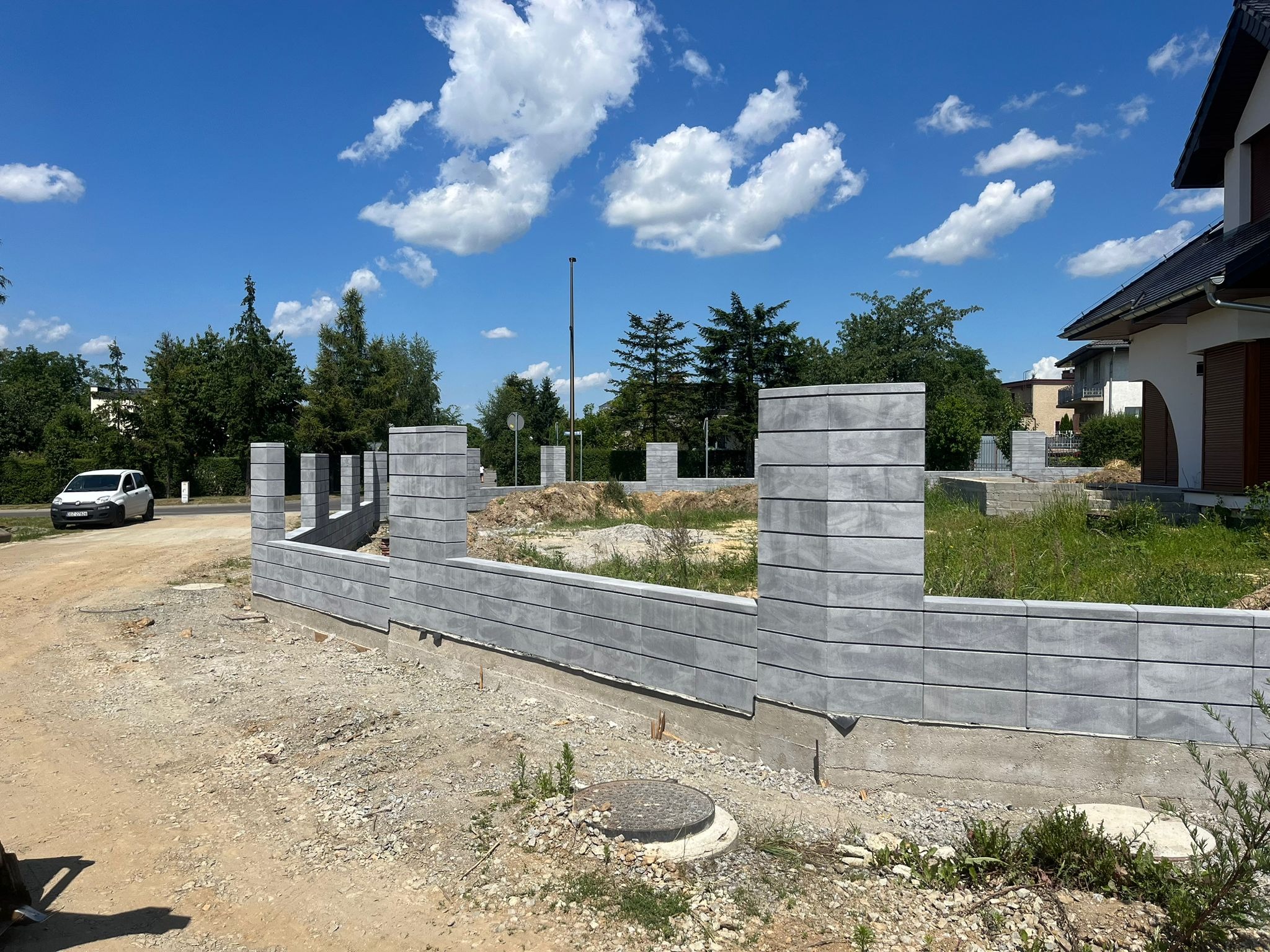 Wznoszenie ogrodzenia betonowego z prefabrykatów modułowych na fundamencie, widoczne słupki i przęsła w trakcie montażu, teren budowy zarośnięty trawą, w tle dom i błękitne niebo.