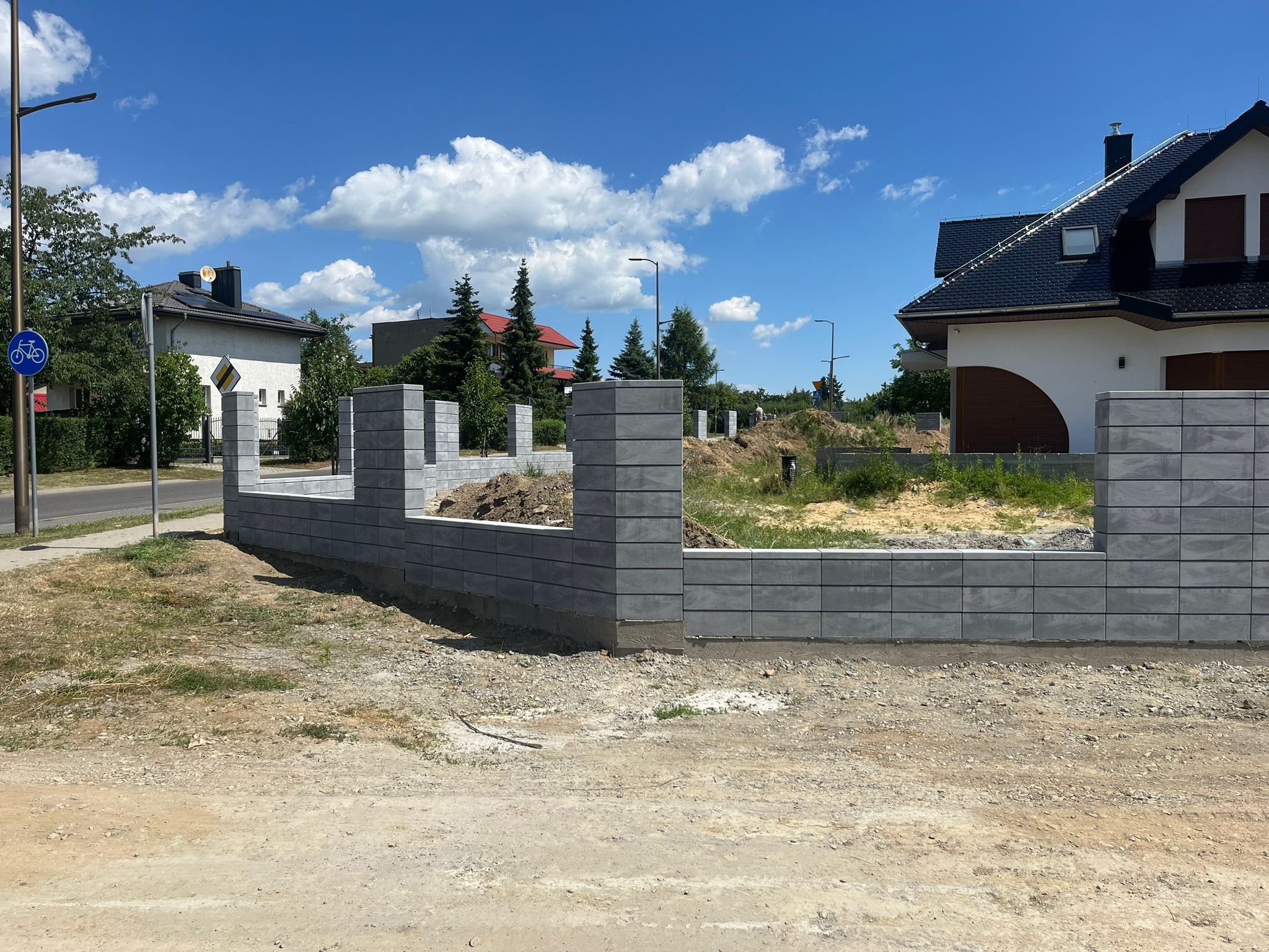 Wznoszony szary płot segmentowy z betonowych bloczków na tle domów jednorodzinnych i błękitnego nieba z chmurami.