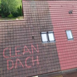 Clean-Dach Mariusz Ciesielski - Częściowo wyczyszczony dach z czerwonej dachówki, widoczne okna dachowe i napis 'CLEAN DACH' na brudnej części dachu, obok zaparkowana przyczepa.