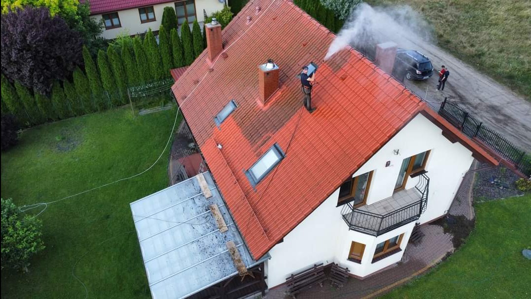 Czerwony dach domu z widocznymi zabrudzeniami czyszczony pod ciśnieniem przez pracownika z użyciem węża, drugi pracownik przy samochodzie, widok z góry.