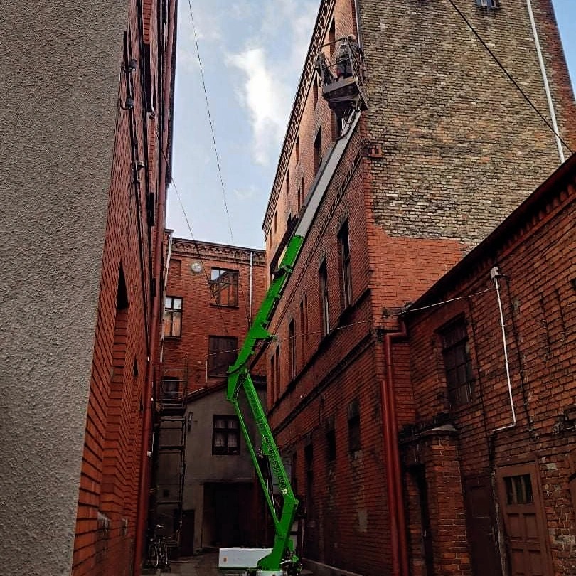 Wąski zaułek między ceglanymi budynkami z podnośnikiem koszowym sięgającym dachu, pracownik w koszu na wysokości, widoczne przewody elektryczne.