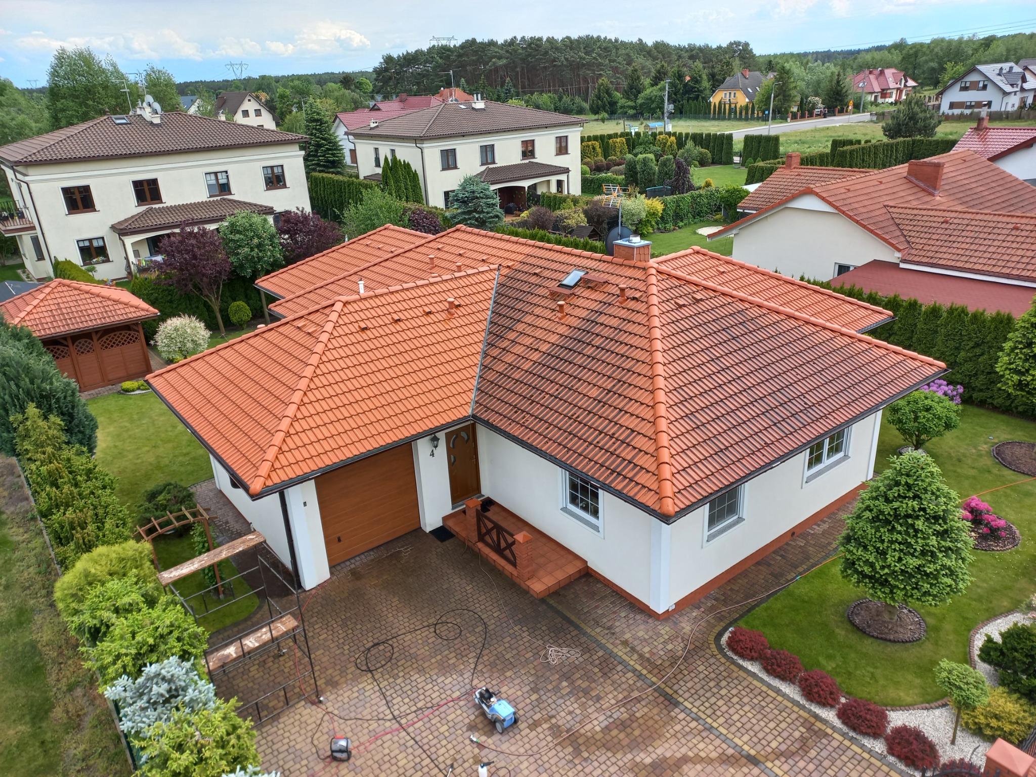 Widok z góry na dom z nową, pomarańczową dachówką. Na podjeździe widoczny sprzęt do czyszczenia, a obok rusztowanie. Zadbane otoczenie z trawnikiem i rabatami.