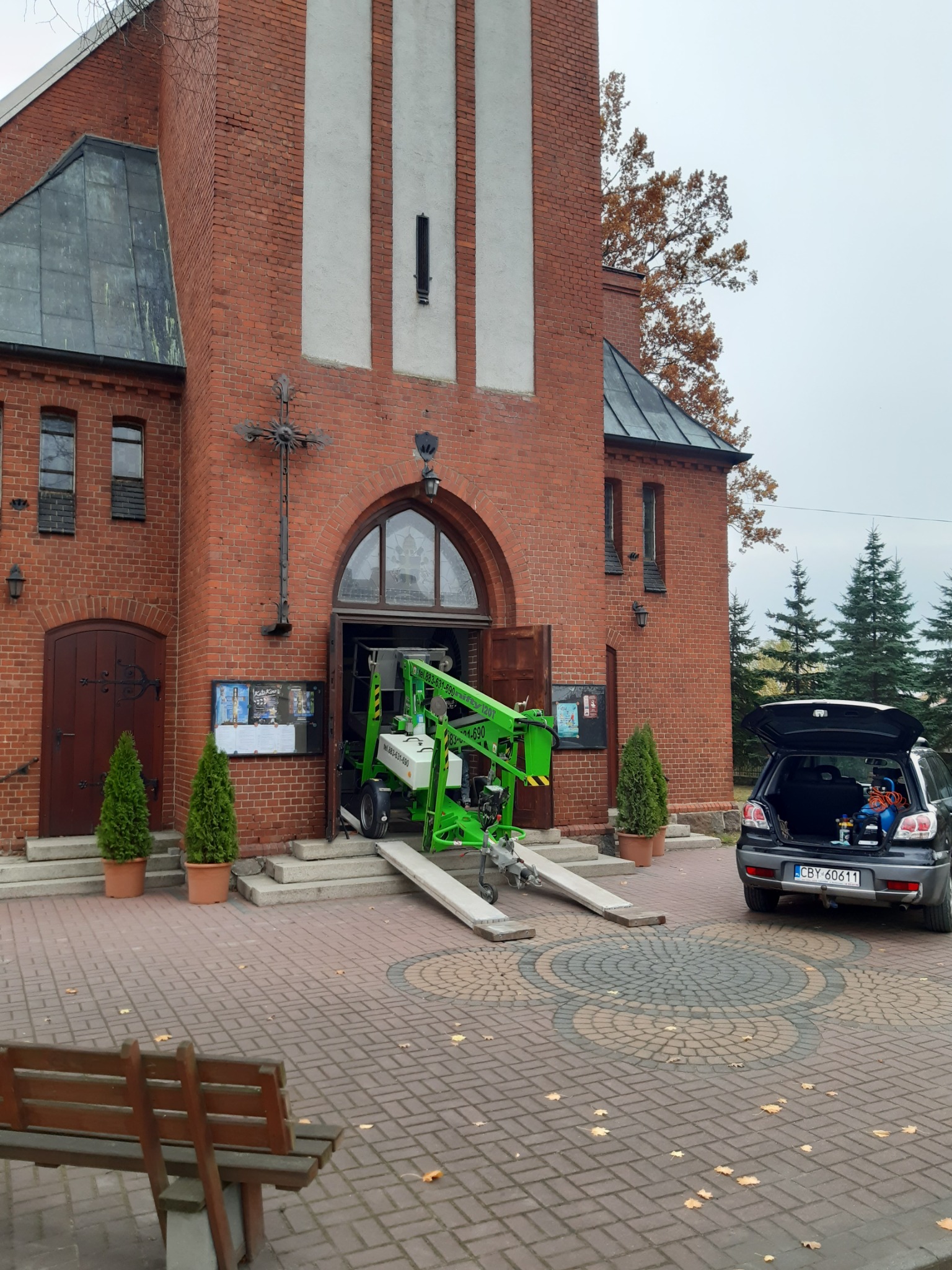 Zielony podnośnik koszowy wjeżdża do wejścia kościoła po rampach, otwarty bagażnik samochodu z narzędziami w tle, jesienne liście na brukowanym placu.