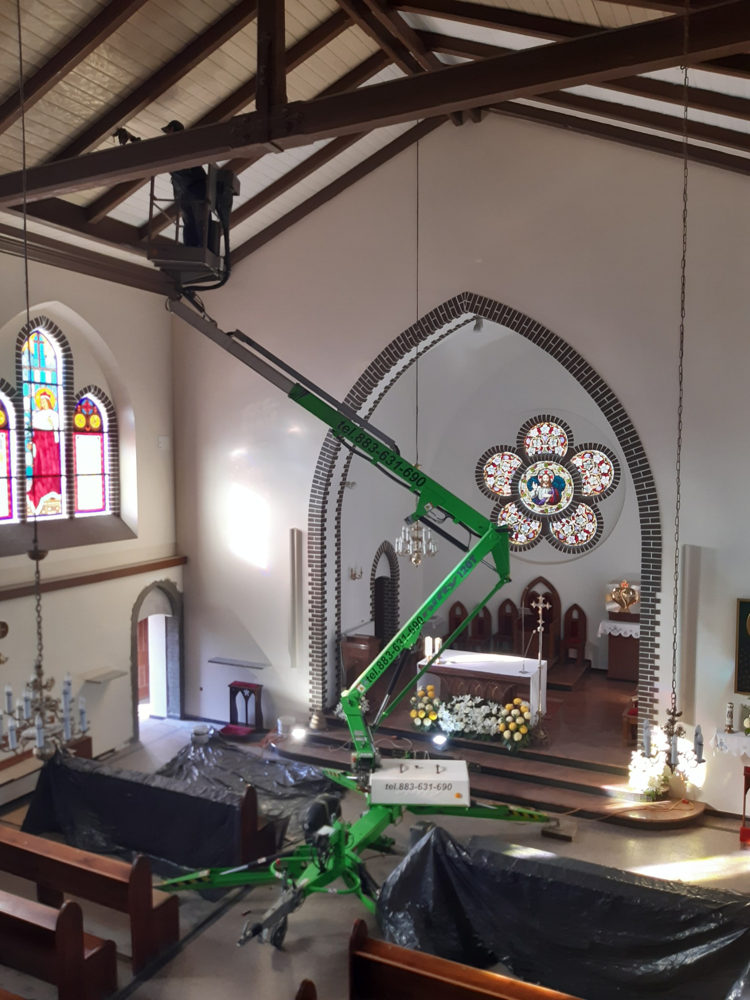 Wnętrze kościoła z podnośnikiem koszowym, pracownik na wysokości przy drewnianej konstrukcji dachu, ławki przykryte folią ochronną, witraże w oknach i nad ołtarzem.