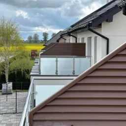 Nowoczesny balkon z balustradą ze szkła i stali nierdzewnej, widok z perspektywy dachu, w tle żółte pole rzepaku i zabudowa jednorodzinna.