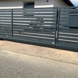 Przesuwna, pozioma brama wjazdowa w kolorze grafitowym z wbudowaną skrzynką na listy, widoczna z zewnątrz posesji, z fragmentem chodnika i ulicy w kadrze.