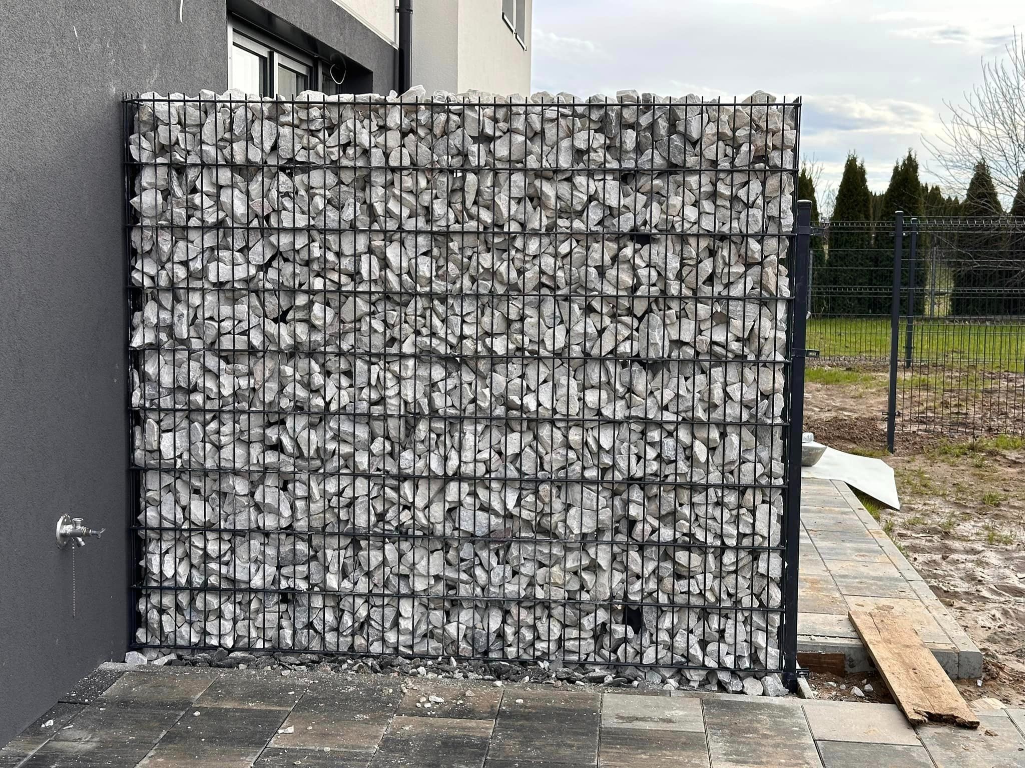 Gabionowa konstrukcja z szarego kamienia w czarnej metalowej siatce, przylegająca do ściany budynku, z widocznym kranem na elewacji i kostką brukową na pierwszym planie.