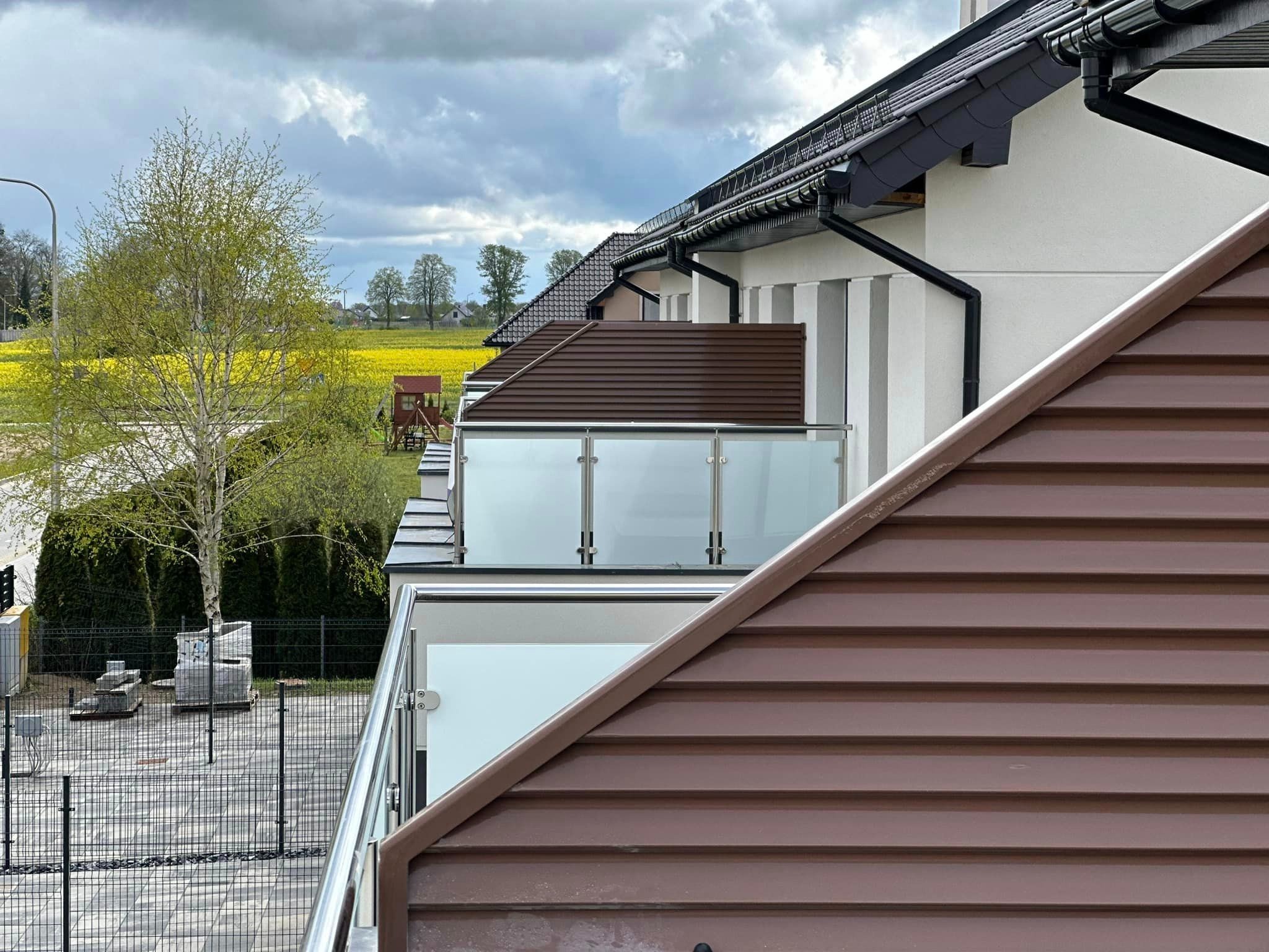 Nowoczesny balkon z balustradą ze szkła i stali nierdzewnej, widok z perspektywy dachu, w tle żółte pole rzepaku i zabudowa jednorodzinna.