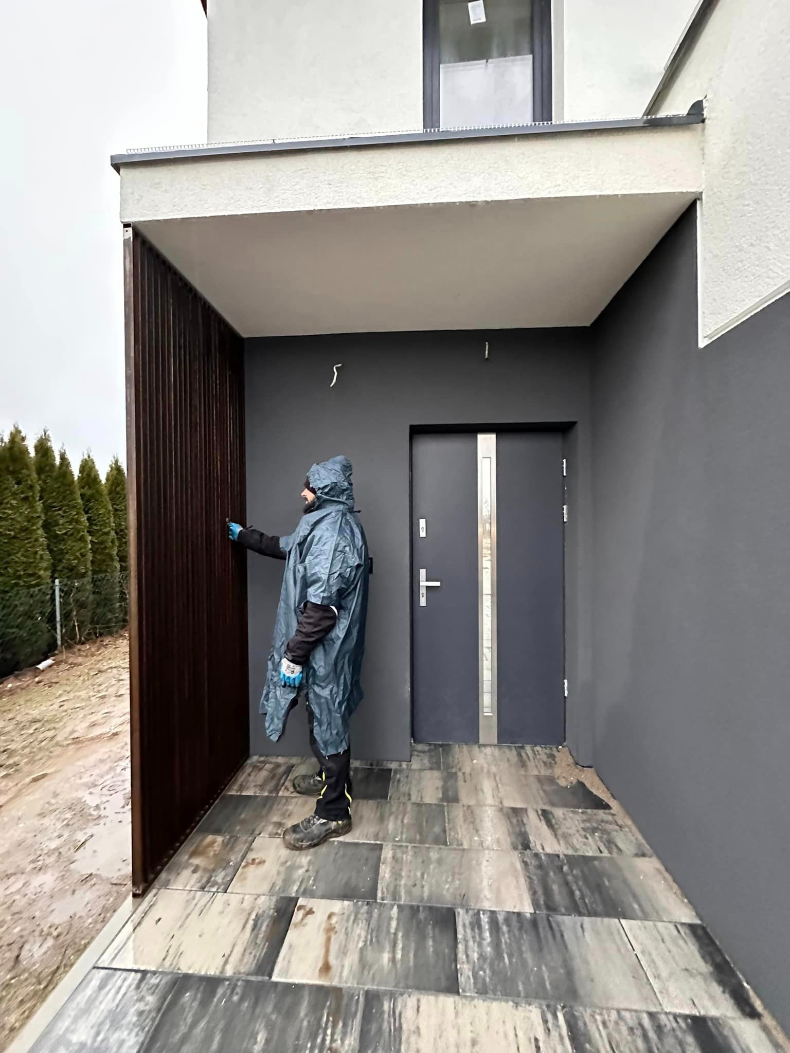 Pracownik w niebieskim płaszczu maluje pionowe, brązowe panele przy wejściu do domu z szarą elewacją i nowoczesnymi drzwiami.