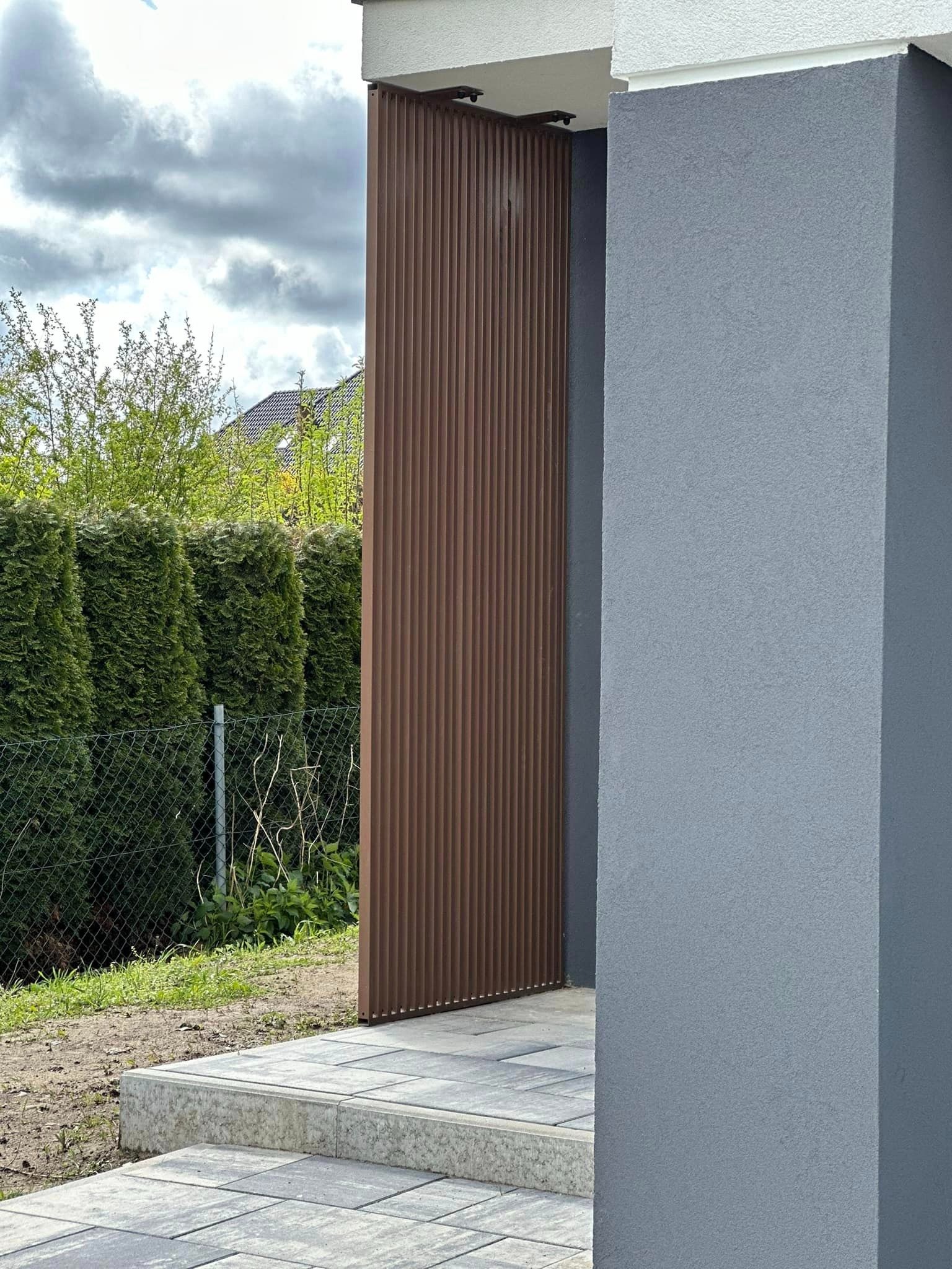 Nowoczesna brama wjazdowa z brązowych paneli pionowych, widoczna z boku, z fragmentem szarej elewacji domu i kostki brukowej, w tle żywopłot z tuj.