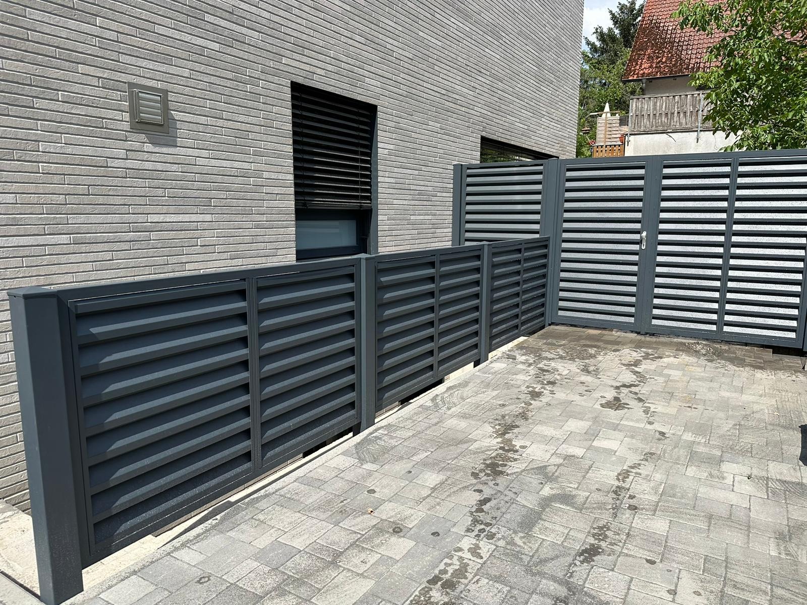 Nowoczesne, poziome, szare przęsła ogrodzeniowe z bramą wjazdową, dopasowane do elewacji budynku z szarej cegły, widoczne z perspektywy chodnika z szarej kostki.
