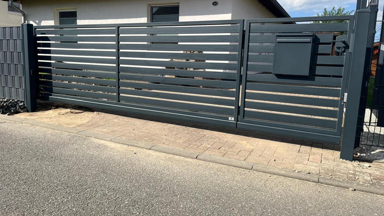 Przesuwna, pozioma brama wjazdowa w kolorze grafitowym z wbudowaną skrzynką na listy, widoczna z zewnątrz posesji, z fragmentem chodnika i ulicy w kadrze.