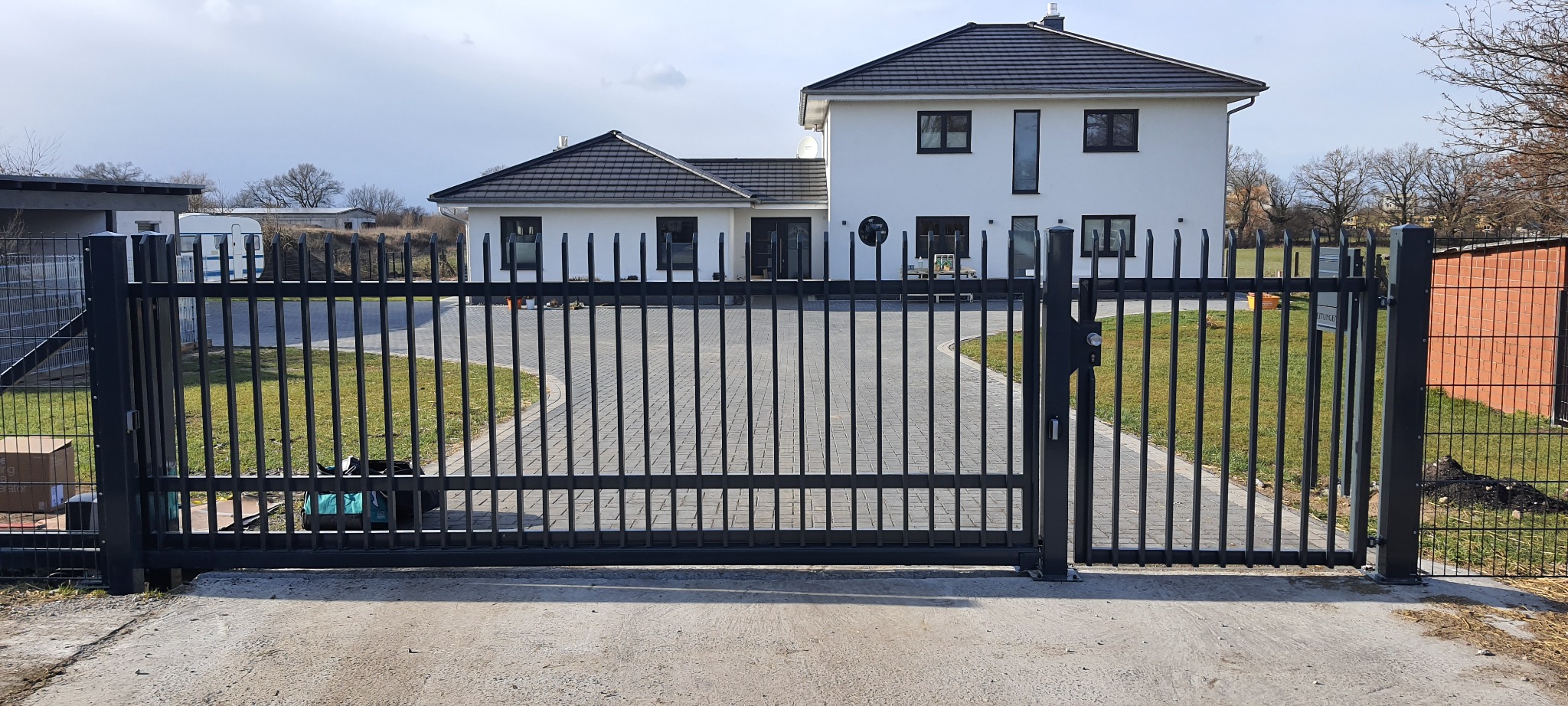 Nowoczesna, czarna brama wjazdowa z pionowymi prętami na posesję z kostki brukowej i białym domem w tle.