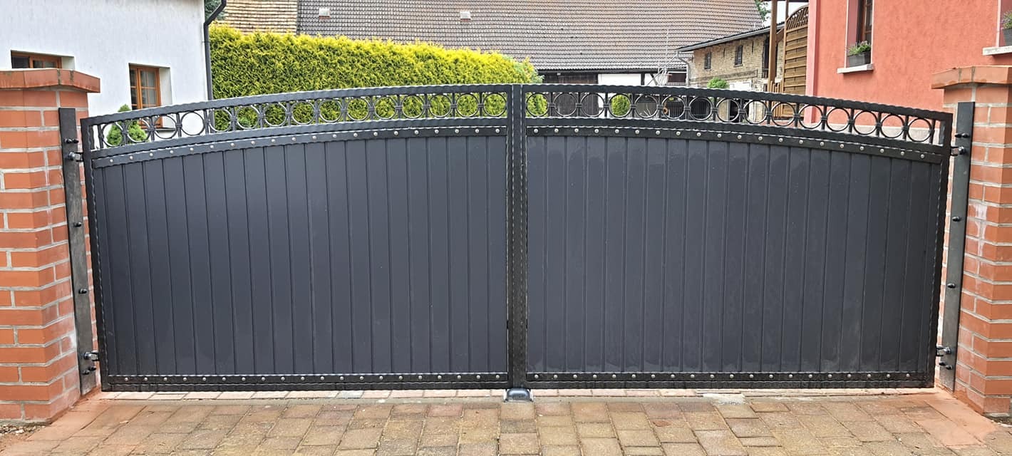 Czarna brama wjazdowa z metalowymi sztachetami i dekoracyjnym, ażurowym wykończeniem w kształcie kół, zamontowana między ceglanymi słupkami na posesji.