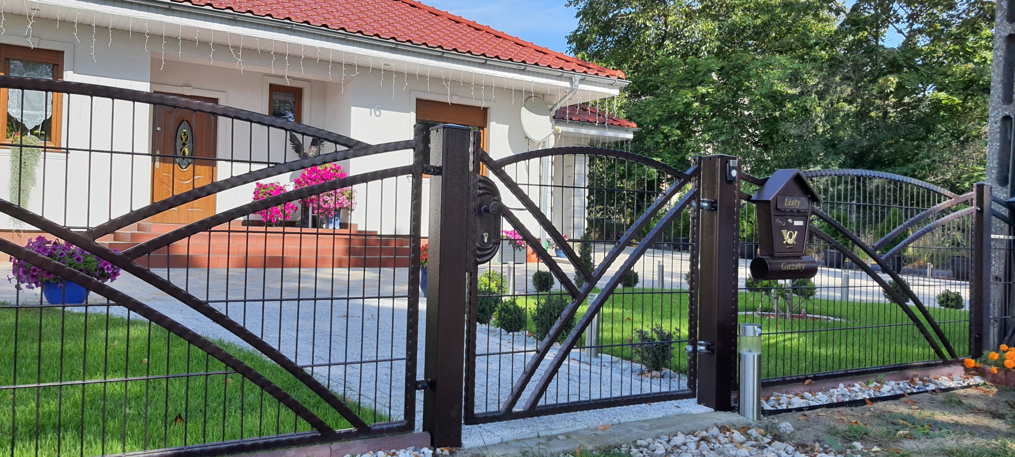 Nowoczesna brama wjazdowa z metaloplastyki, brązowa, z dekoracyjnym łukiem i skrzynką na listy w stylu retro, na tle domu z czerwoną dachówką i zielonego trawnika.