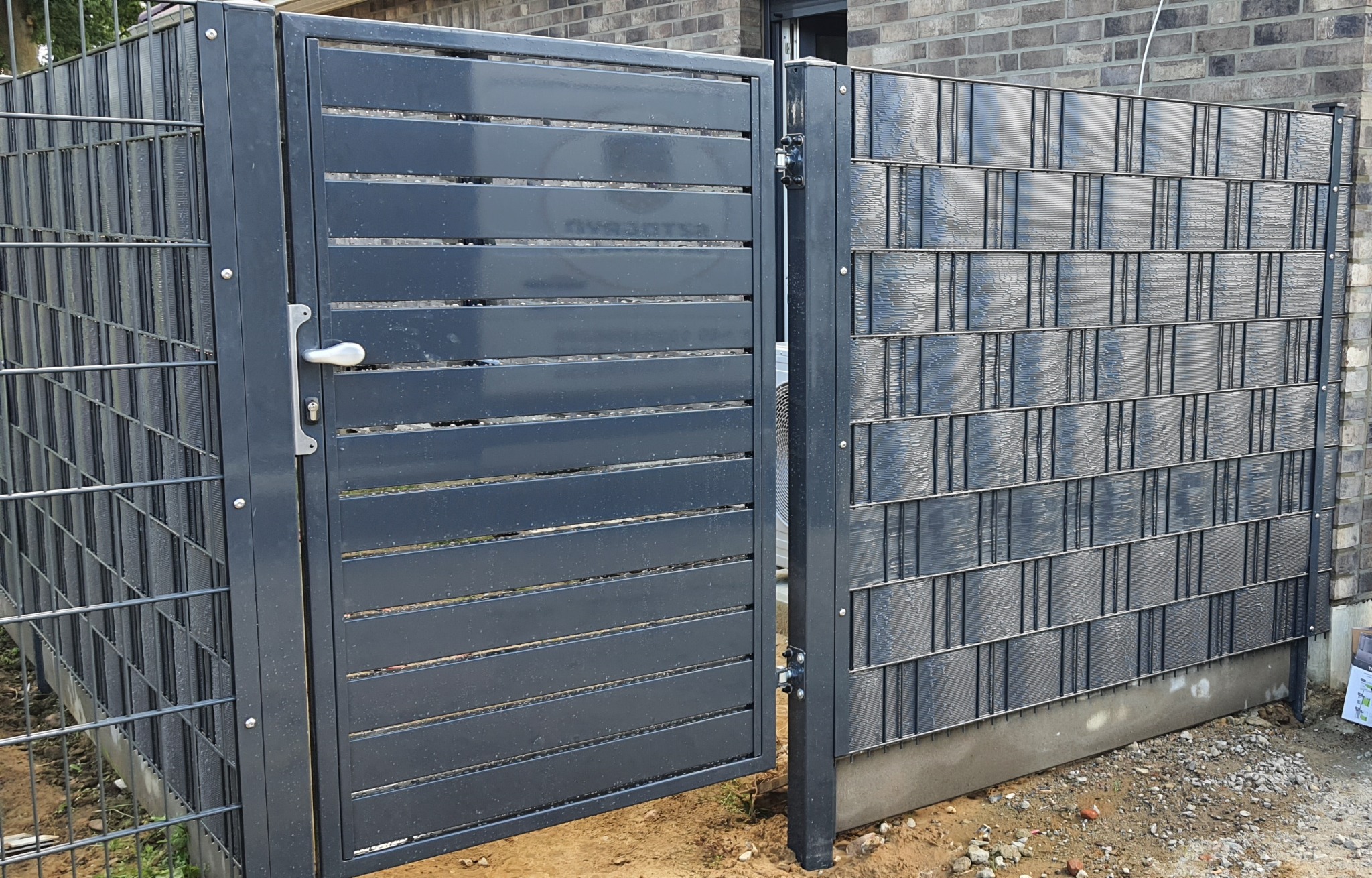 Otwarta, szara brama z poziomych listew, wkomponowana w panelowe ogrodzenie z siatki, z widocznym betonowym fundamentem i fragmentem ceglanego budynku w tle.
