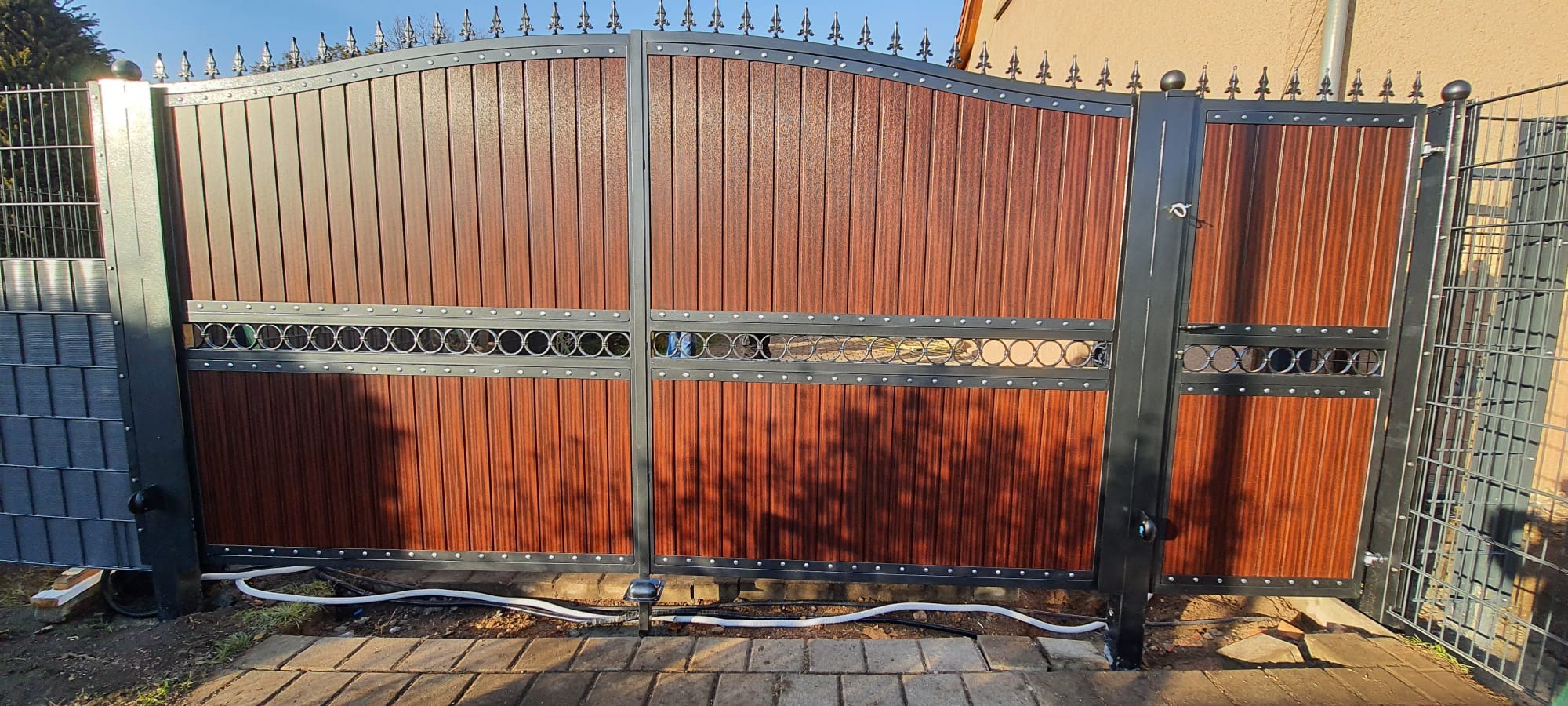 Dwuskrzydłowa brama wjazdowa z brązowymi panelami imitującymi drewno, zdobiona metalowymi elementami z motywem kół, zamontowana na posesji z widocznymi kablami i kostką brukową.