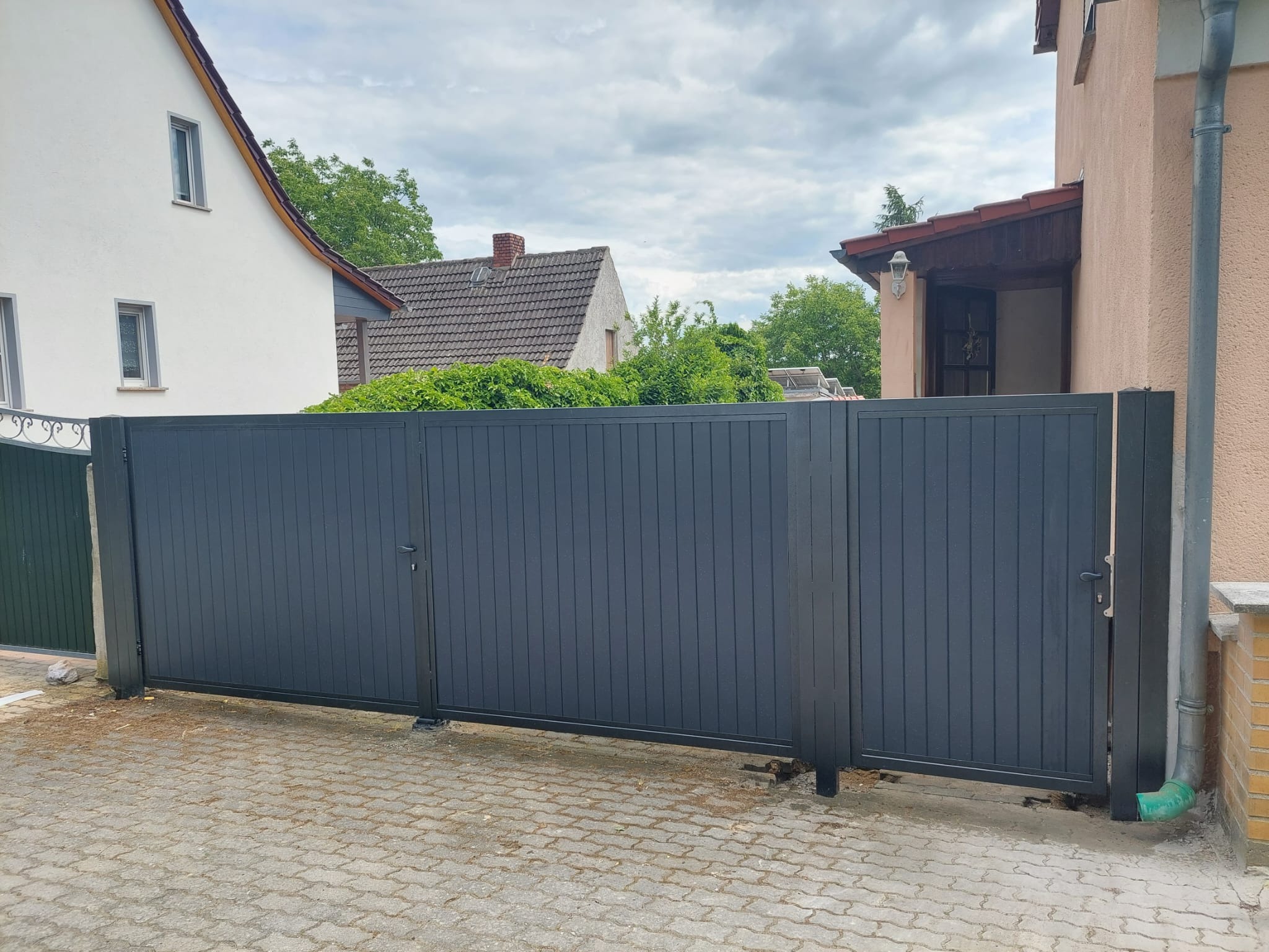 Nowoczesna, przesuwna brama wjazdowa w kolorze grafitowym, z pionowymi panelami, zamontowana przed domem z kostki brukowej, widoczny fragment rynny spustowej.