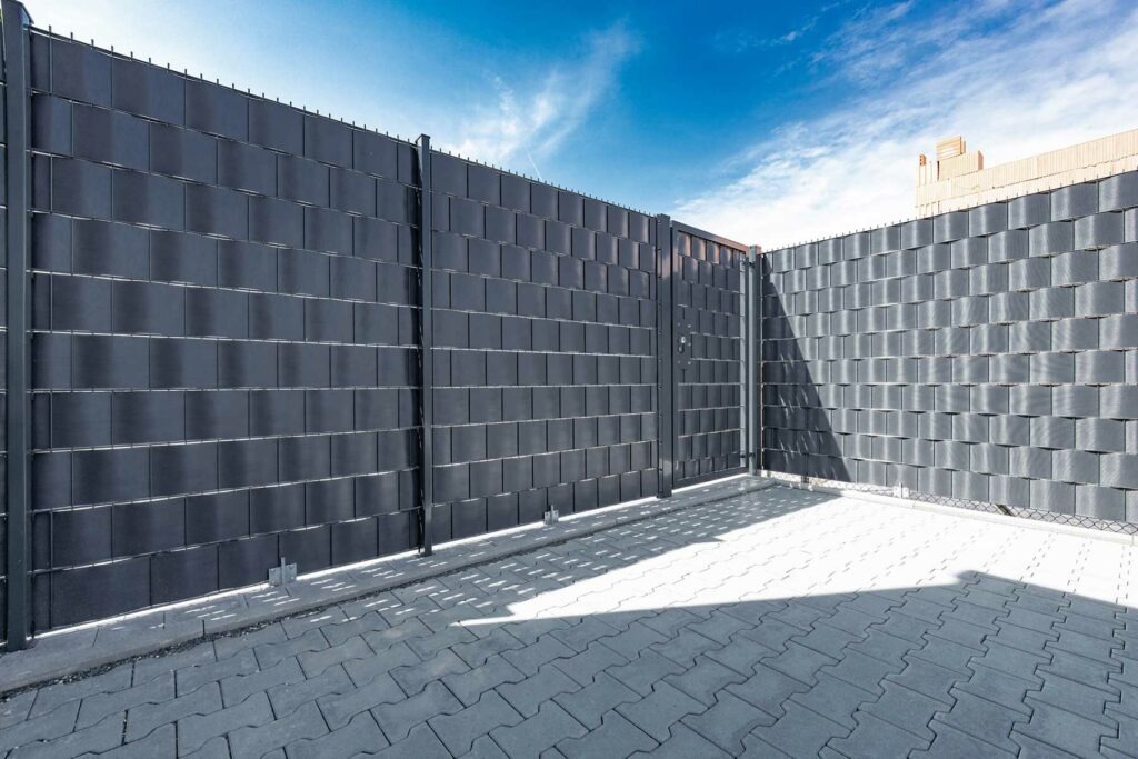 Grafitowe ogrodzenie z paneli kompozytowych z bramą wjazdową, widziane w słoneczny dzień na tle błękitnego nieba z delikatnymi chmurami.