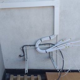 Pro-Split - Białe kable zabezpieczone czarnymi opaskami, prowadzone wzdłuż ściany i przymocowane do metalowego wspornika, z widocznymi końcówkami przewodów oraz fragmentem drewnianej palety na podłodze.